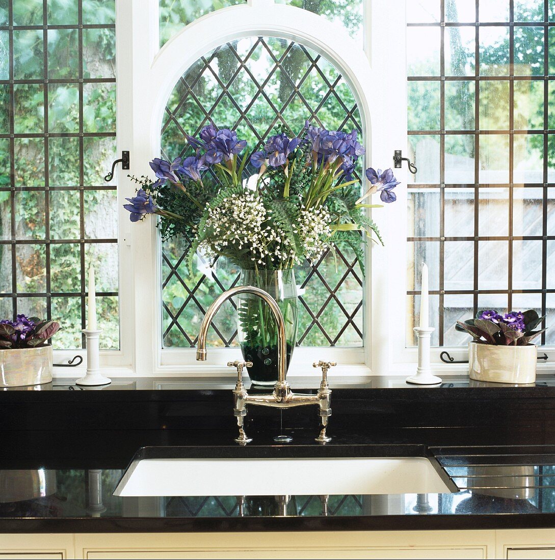 Blaue Iris vor vergittertem Rundbogenfenster über altmodischem Wasserhahn in schwarzer Küchenarbeitsfläche