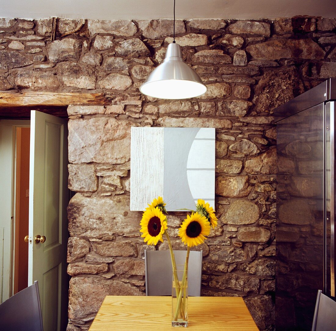Modernes Esszimmer mit Edelstahlkühlschrank im Kontrast zu Natursteinmauer und Sonnenblumenstrauss auf Holztisch