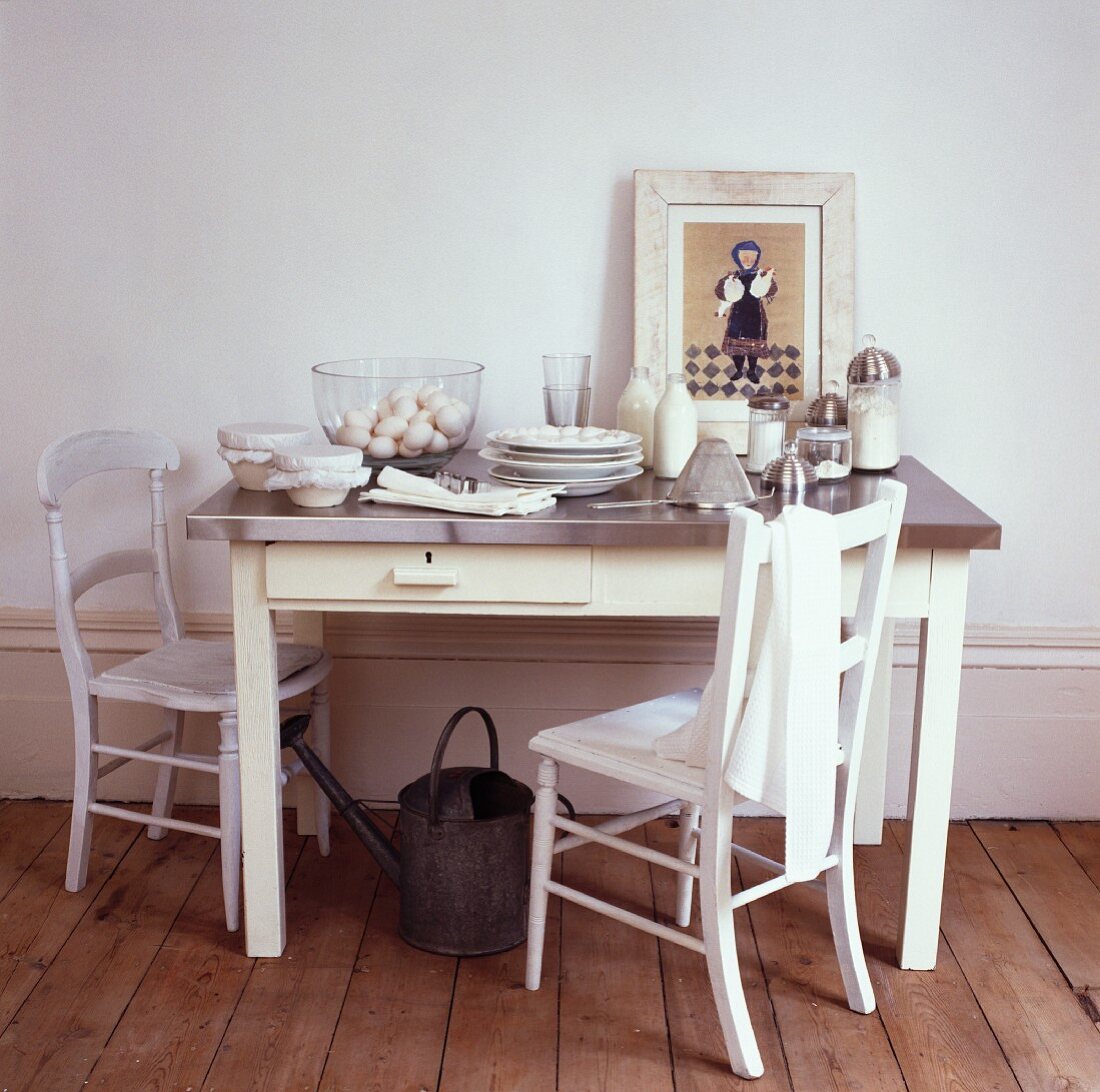 Mit Backutensilien vollgestellte Edelstahlplatte auf cremefarbenem Holztisch und einfachen Stühlen auf unlackiertem Dielenboden