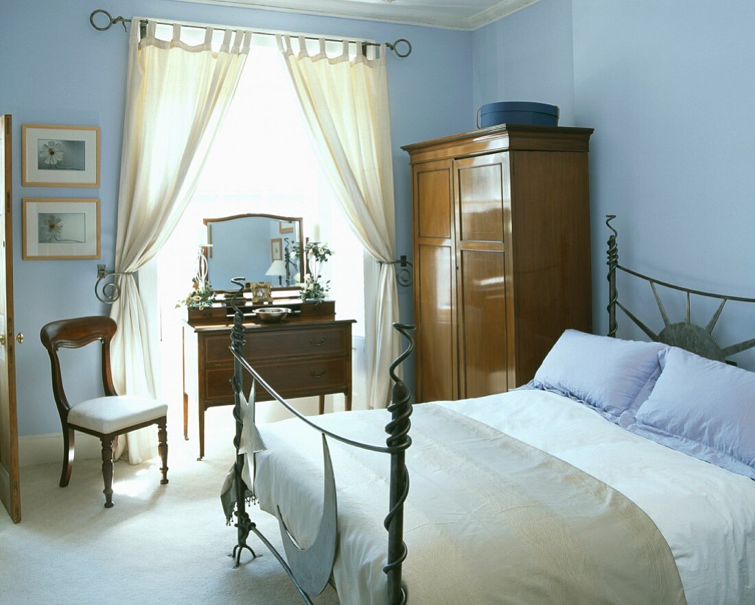 Lichtblaues Schlafzimmer mit modernem Metallbett neben Biedermeier-Stuhl, Schrank und Frisierkommode vor dem Fenster