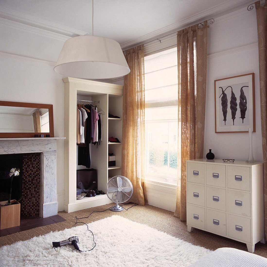 Haartrockner und Ventilator auf dem Boden in einem Schlafzimmer mit weisser Kommode und beigefarbenen Gardinen