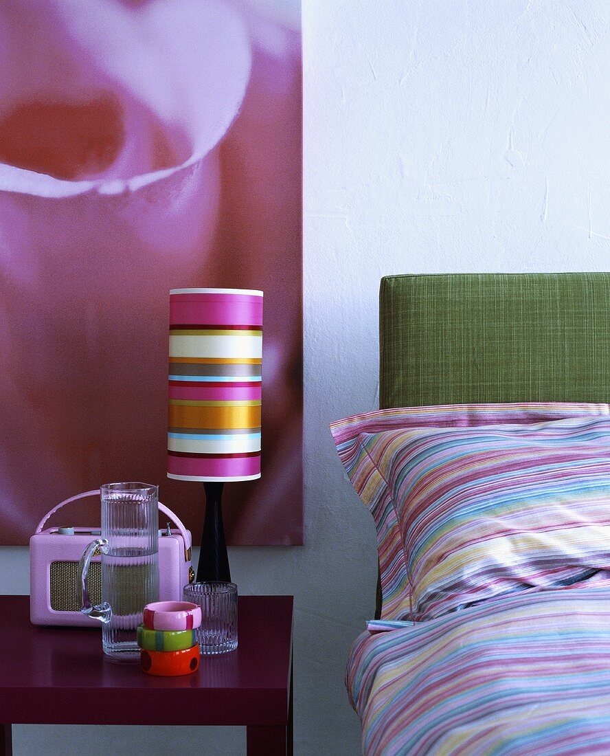 Pinkfarbenes Roberts Radio und eine Lampe mit gestreiftem Schirm auf dem Tisch neben einem Bett mit gestreifter Bettwäsche