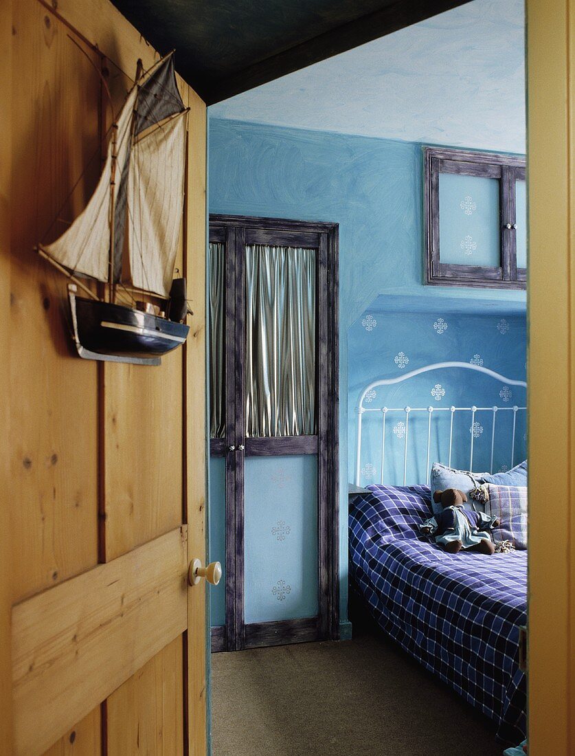 Blick durch Kiefernholztür in Kinderschlafzimmer auf weisses Metallbett mit Teddy und Muster in Schablonentechnik auf hellblauer Wand