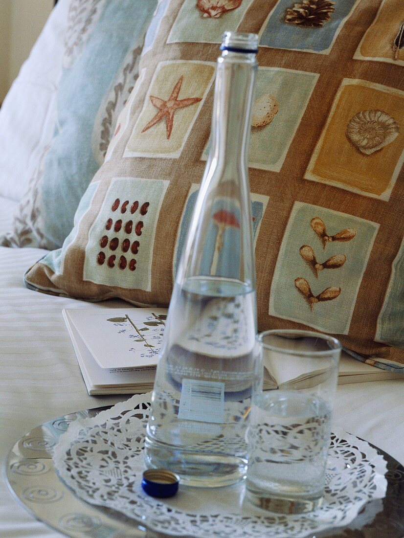Tablett mit Wasserglas und Wasserflasche auf dem Bett mit gemusterten Kissen