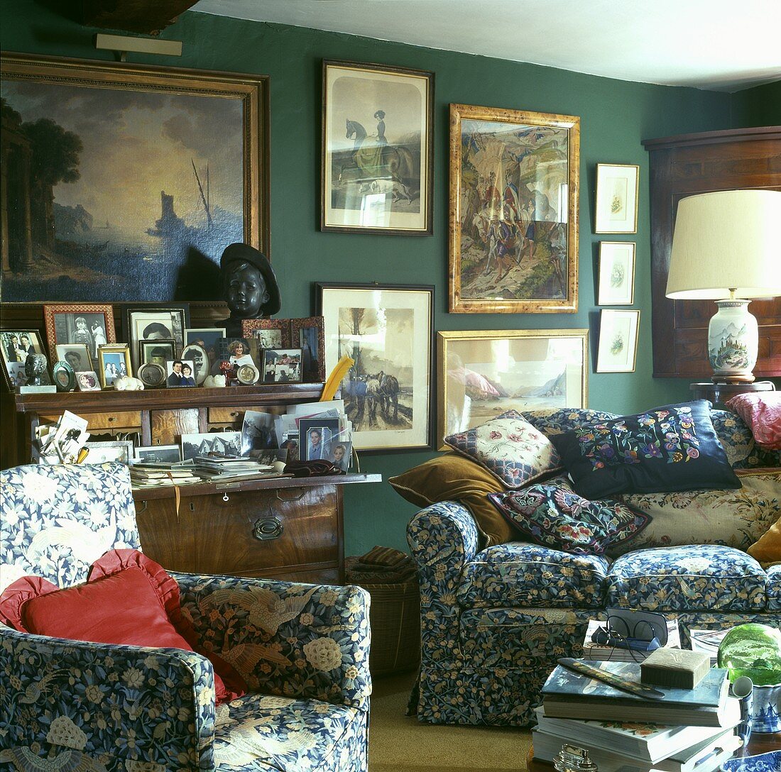 Polstermöbel mit floralem Muster und Kissen vor Sammlung von Gemälden an der Wand und kleinen Fotos auf Sekretär