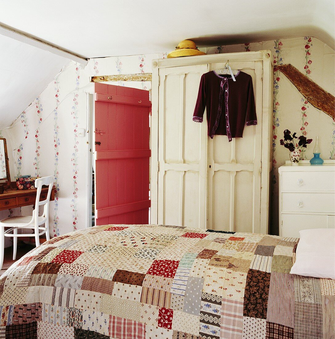 Romantisches Schlafzimmer unter dem Dach mit Patchworkdecke auf dem Bett, gemalten Blumengirlanden an der Wand und rosafarbener Holztür