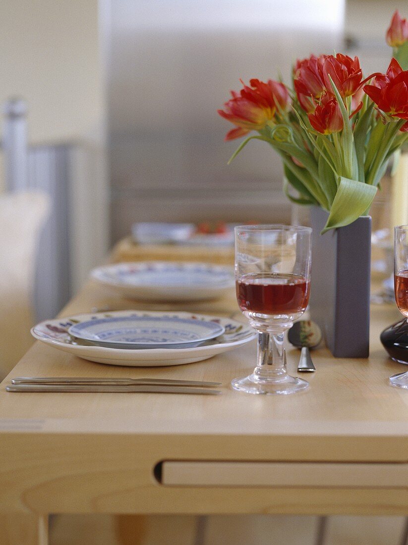 Nahaufnahme von roten Tulpen in grauer Vase auf hellem Holztisch mit Gedeck und einem Glas Rotwein