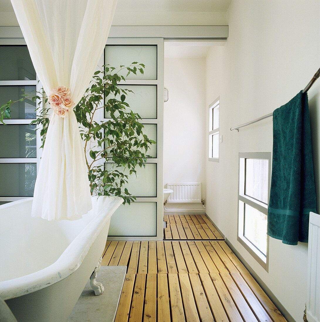 Holzdielenboden und Schiebetüren aus Glas dominieren das moderne Bad mit einer freistehenden Badewanne, über die ein weißer Vorhang mit einem Bund aus rosa Rosen zusammengebunden wurde
