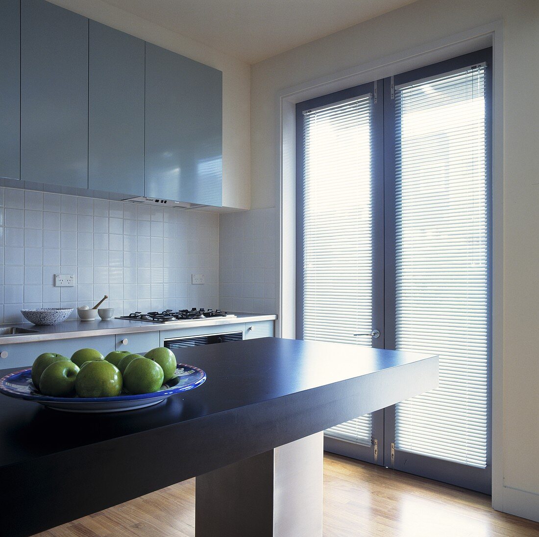 Ein Teller mit Äpfeln steht auf einer grauen freistehenden Arbeitsplatte in einer hellgrauen Küche mit einem Flügelfenster, das zu einer Terrasse führen und an dem Jalousien angebracht sind