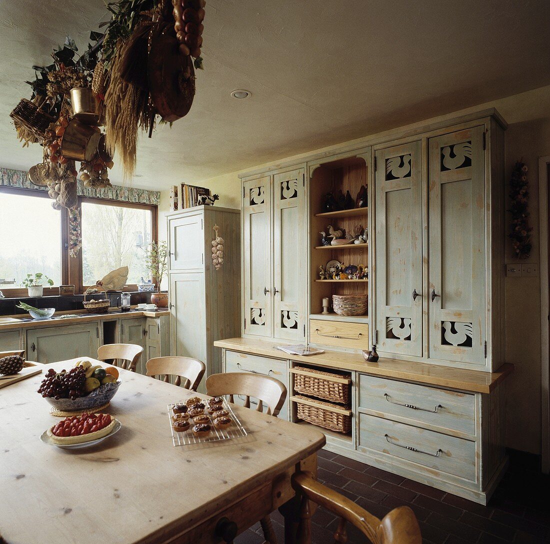 Geschnitzte grau lackierte Wandschränke stehen in einer ländlichen Küche mit einem alten Kiefern Tisch und Stühlen
