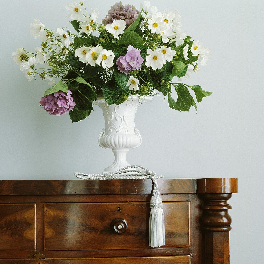 Nahaufnahme eines weiss-violetten Blumenstrausses in weisser Amphorenvase auf antiker Holzkommode