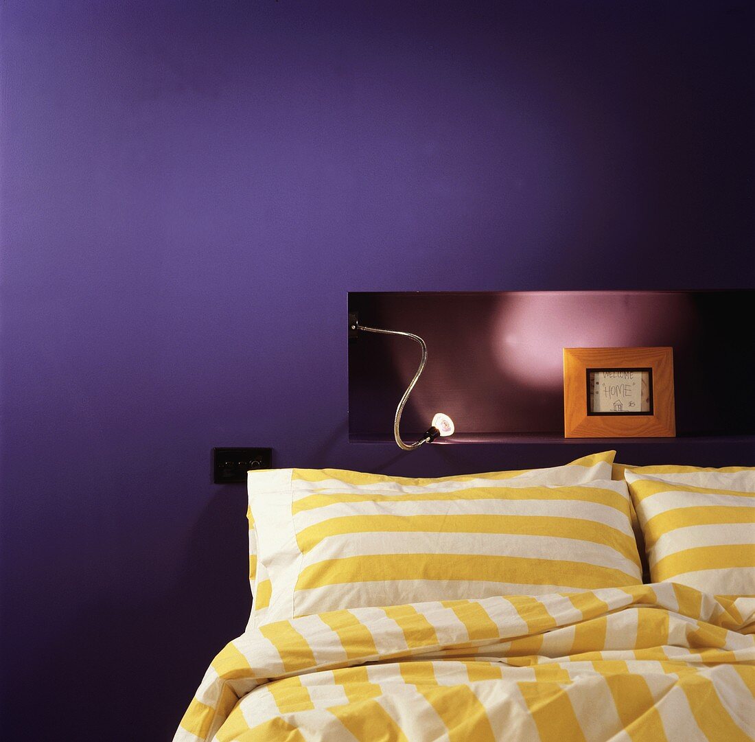 Gelbweiss gestreifte Bettwäsche vor beleuchteter Nische in lilafarbener Wand mit gerahmtem Bild