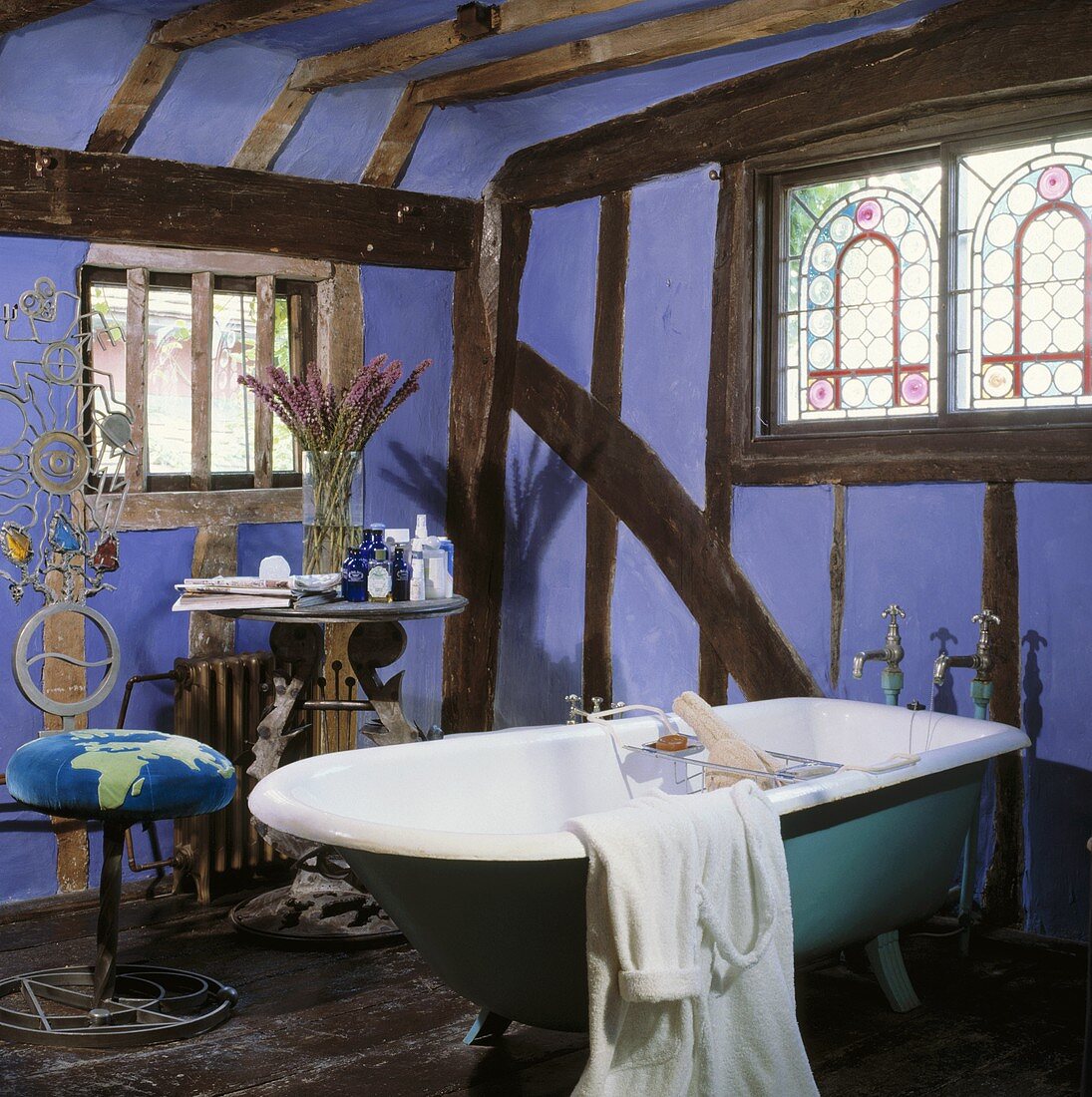 Altes Fachwerk mit lavendelfarbener Ausfachung und bunten Bleiglasfenstern über freistehender türkisfarbener Badewanne und Lyra-Tisch
