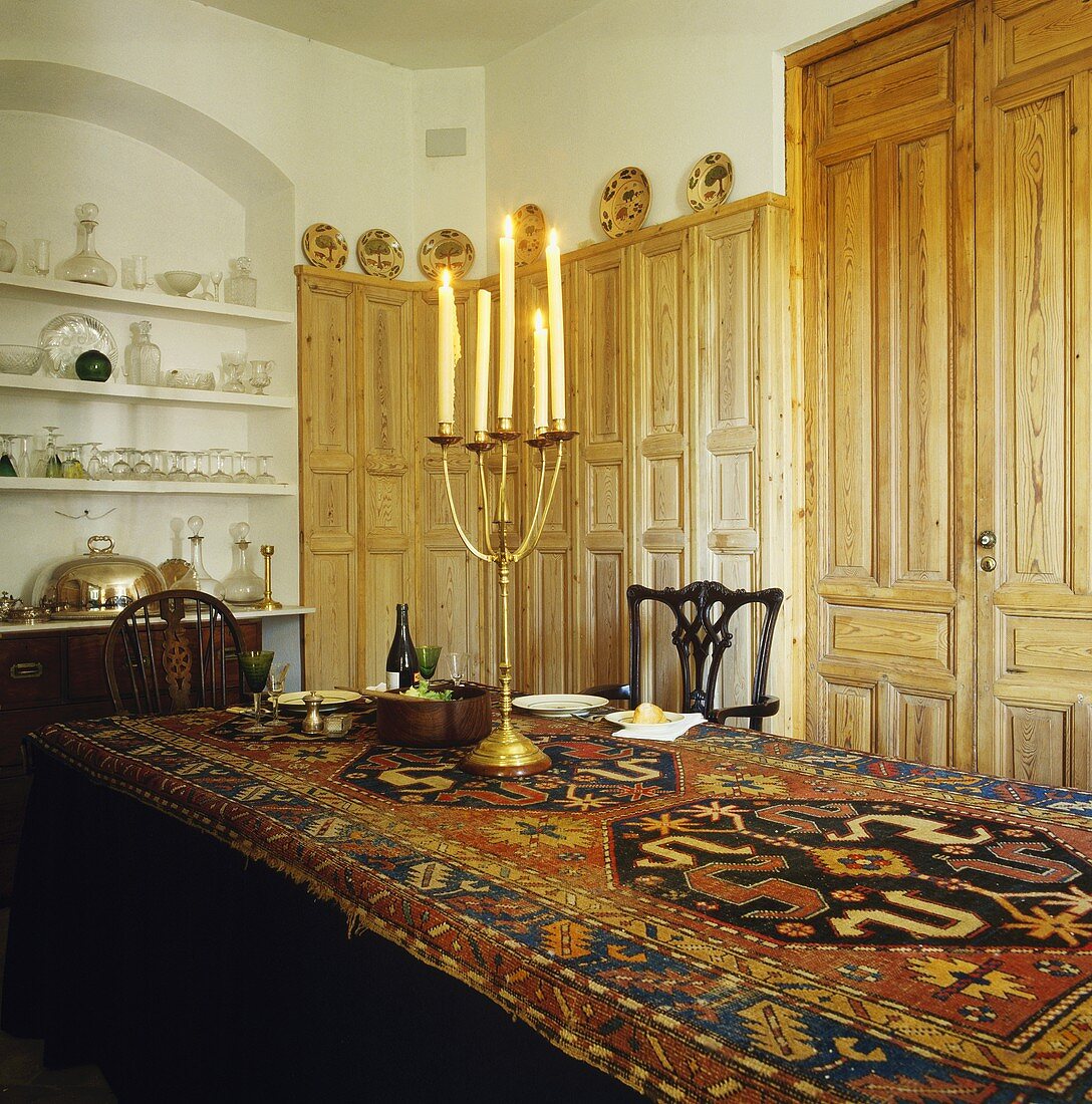 Gedeckte Tafel mit altem Kelim als Tischdecke, Messing-Kerzenleuchter und antiken Holzstühlen vor Wandvertäfelung und Wandschrank aus Kiefer