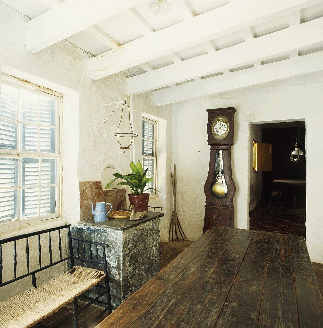 Esszimmer in altem, bäuerlichen Landhaus mit weissen Holzbalken, geschlossenen Fensterläden und antiker Standuhr neben Tür