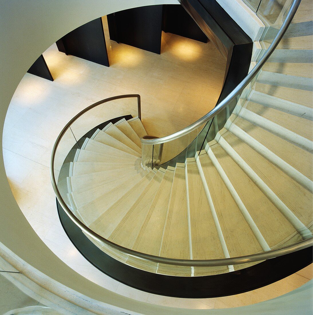 Blick von oben auf moderne geschwungene Treppe mit Geländer aus Glas und hellen Steinstufen
