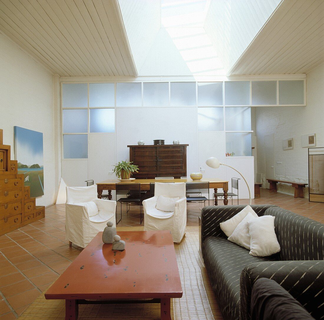 Modernes, loftartiges Wohnzimmer mit rostrotem Couchtisch und Sesseln mit weissen Hussen vor Trennwand mit Licht durch satiniertes Glas