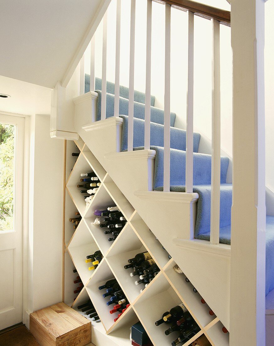 Weinflaschen in auf der Spitze stehendem Würfelregal unter weisser Holztreppe mit hellblauem Teppich