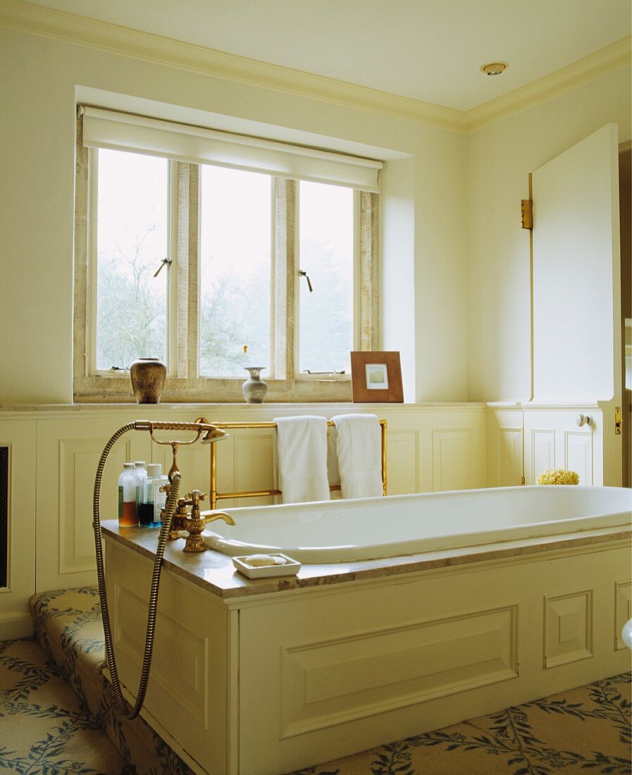 Badewanne mit cremefarbener Holzverkleidung, Granitumrahmung und altmodischer Messingarmatur in der Mitte eines hellen Badezimmers