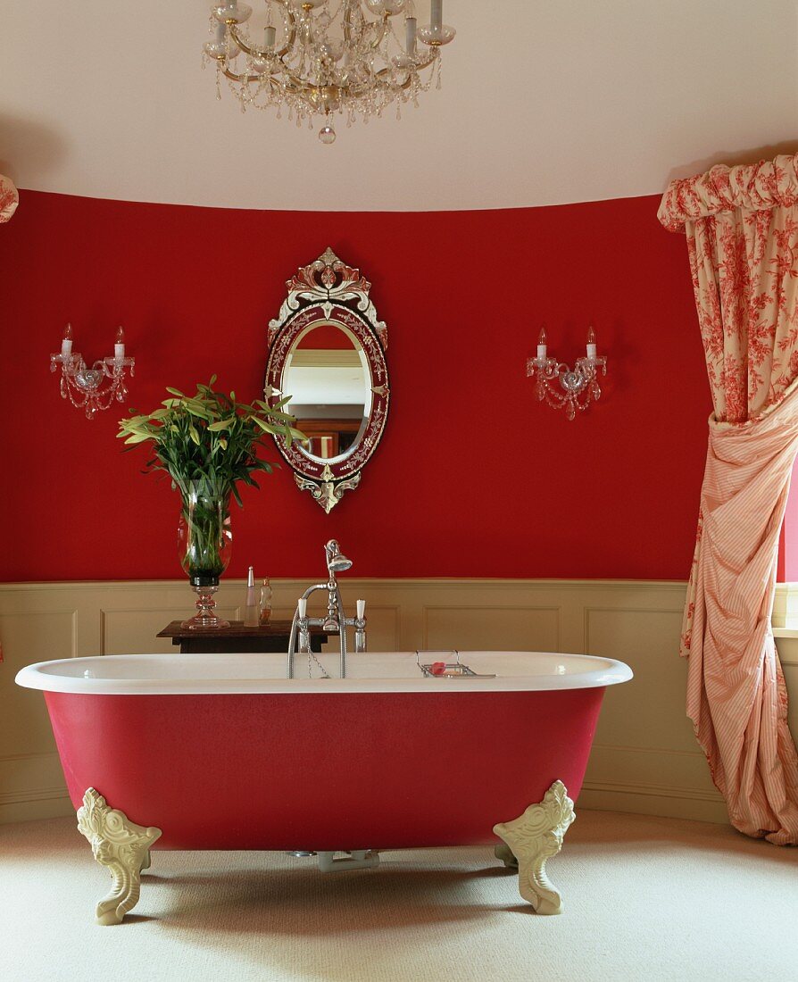 Ovaler, venezianischer Spiegel und Kristallleuchter an roter Wand hinter freistehender Badewanne, himbeerfarben mit weissen Klauenfüssen