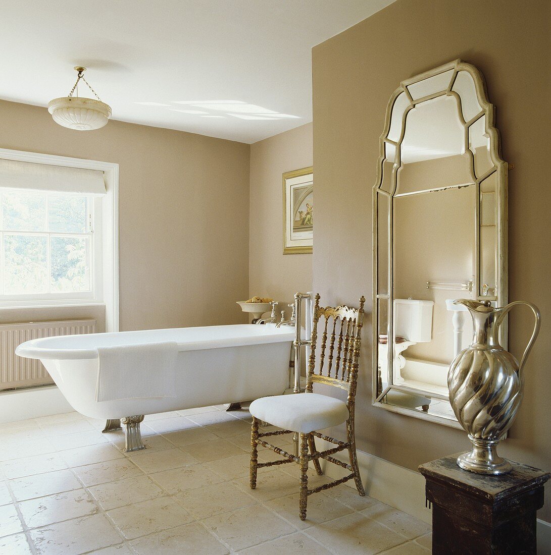 Ein antiker vergoldeter Stuhl vor einem großen Wandspiegel in einem hellen Badezimmer mit frei stehender Badewanne