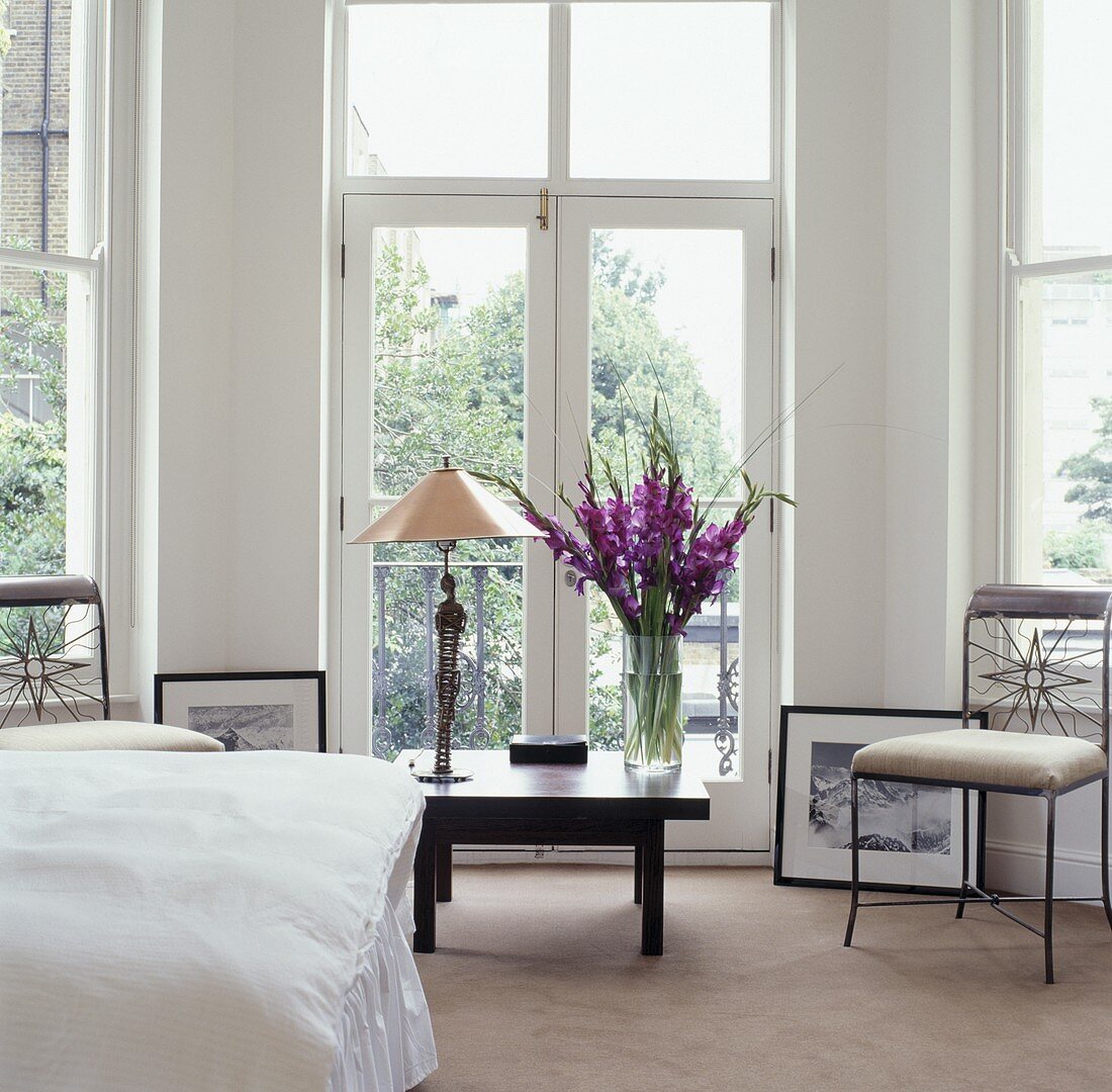 Eine Tischlampe und lila Gladiolen auf einem niedrigen Tisch vor dem französischen Fenster in einem weissen zeitgenössischen Schlafzimmer