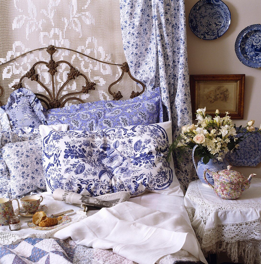 Nahaufnahme von einem schmiedeeisernen Bett mit blau-weiss gemusterten Kissen, Spitzen- und Baumwollvorhang hinter dem Bett und Teekanne auf dem Nachttisch