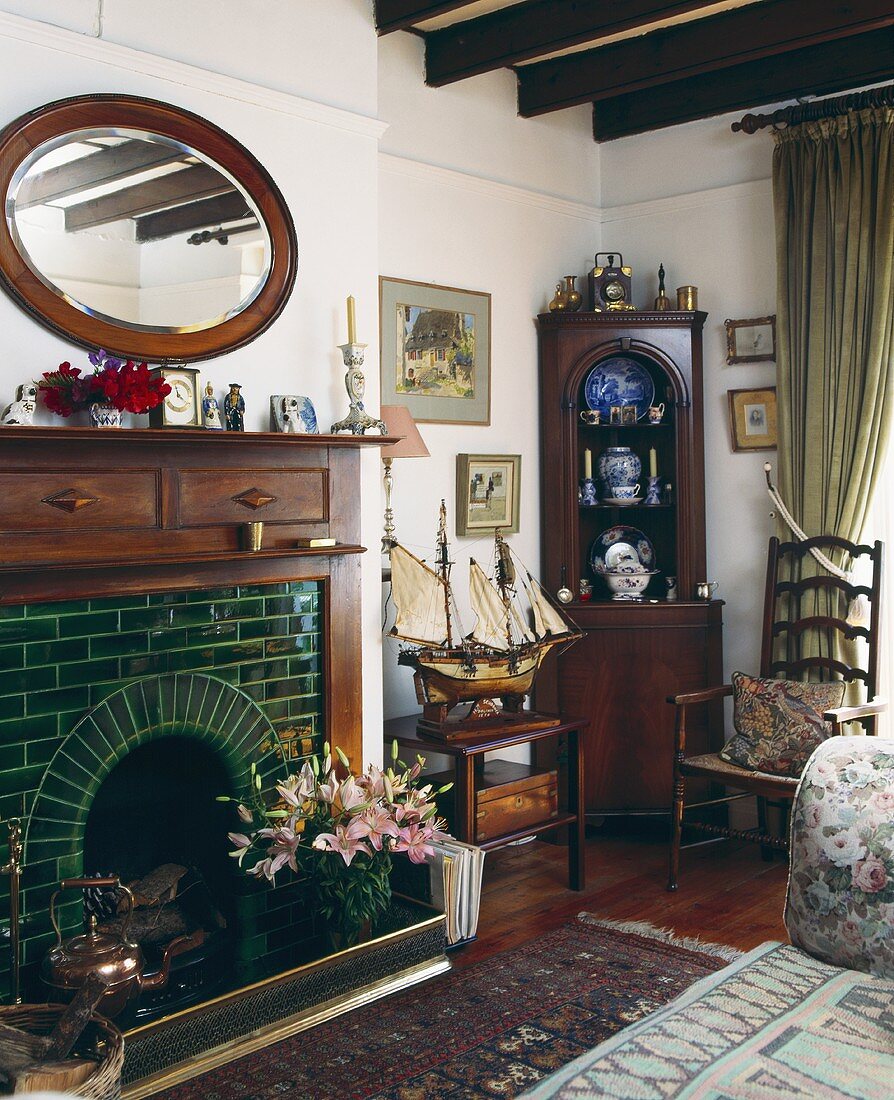 Wohnzimmer mit grün gefliestem Kamin darüber ovaler Spiegel mit Holzrahmen und antiker Eckschrank