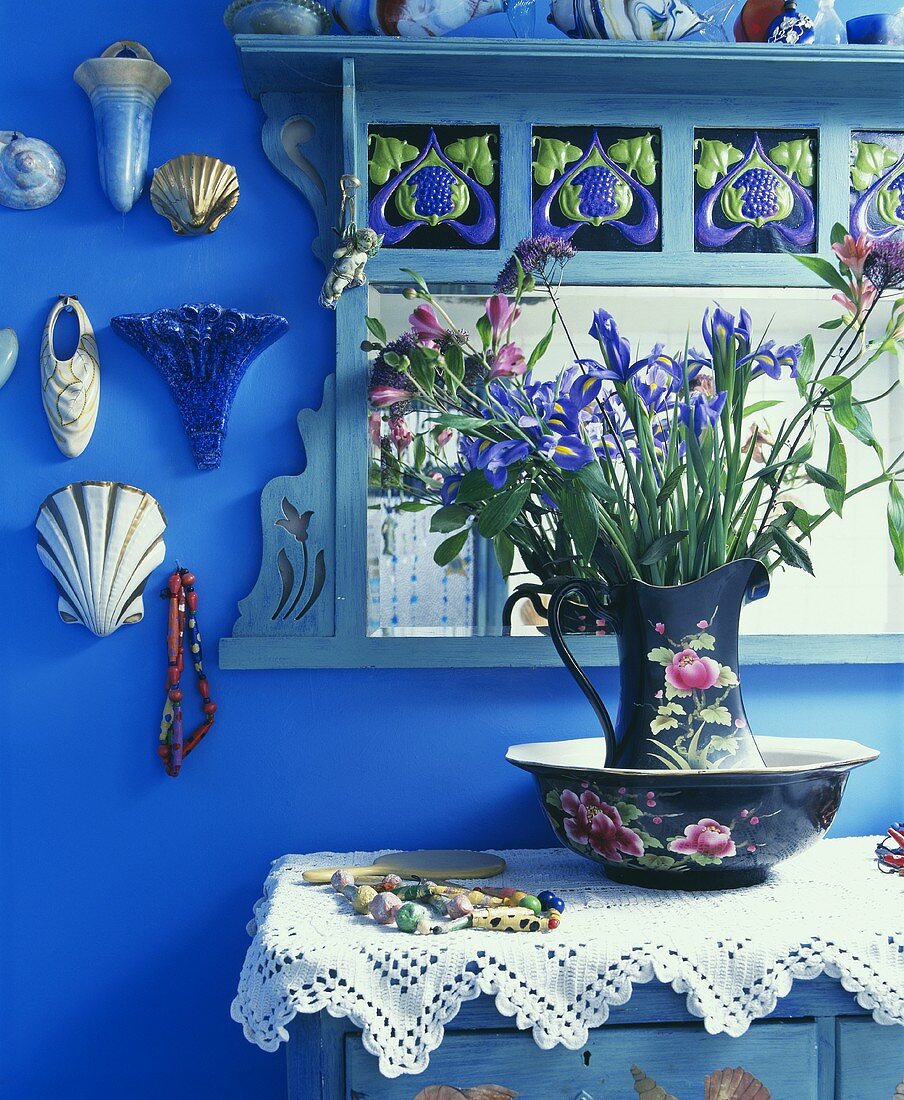 Bunter Frühlingsstrauss im Wasserkrug mit Schüssel auf Spitzendecke vor blauen Wand und Schmuckmotiven aus Porzellan