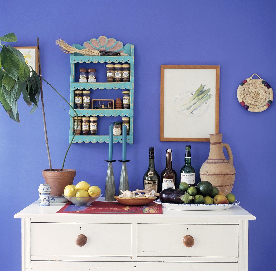 weiße Holzkommode mit Weinflaschen und Schalen mit Obst vor blauer Wand