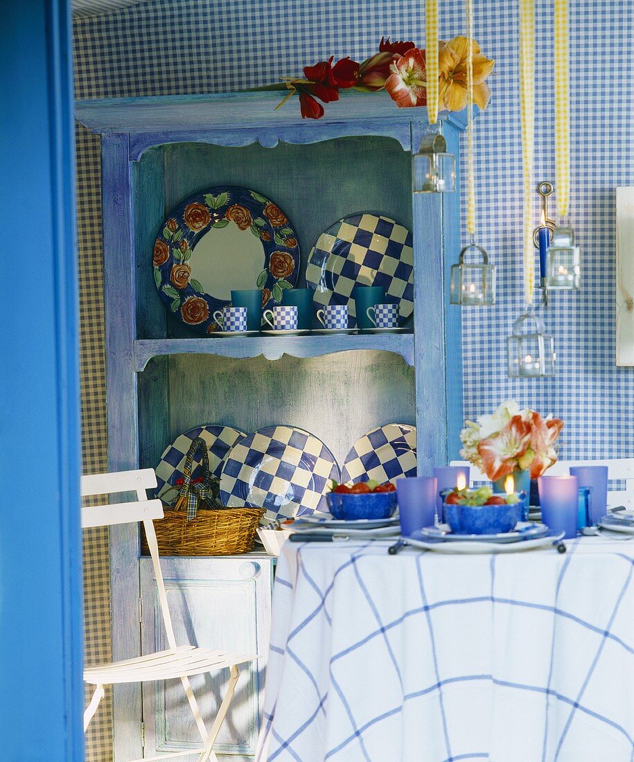 Gedeckter Tisch mit mit blauen Gläsern vor bäuerlichem Regalschrank und Karomuster auf Tapete