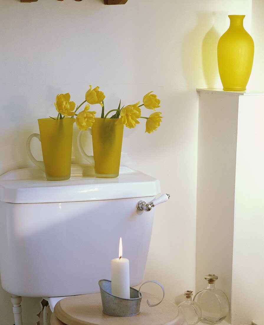 Gelbe Tulpen in gelben Glaskrügen auf Spülkasten und brennende Kerze auf Toilettensitzgarnitur