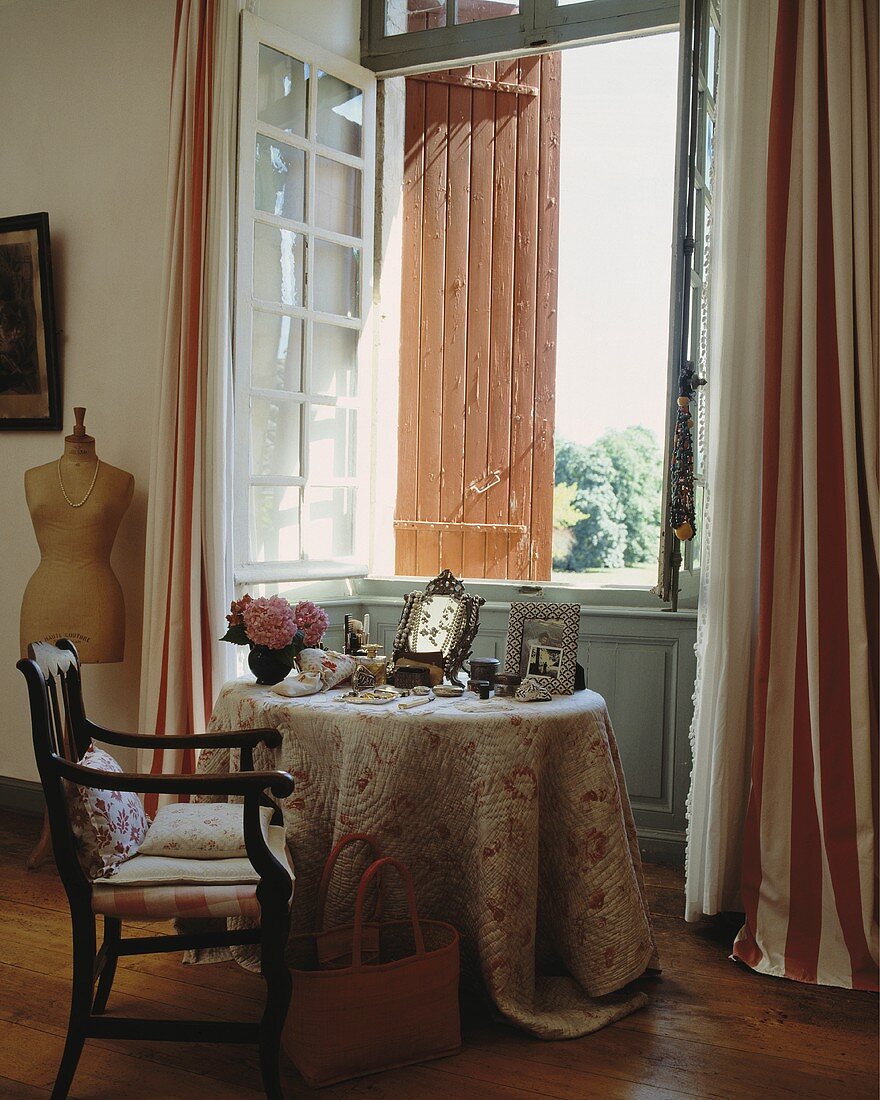 Sitzplatz vor offenem Fenster mit Fensterläden und gestreiften Vorhängen im Schlafzimmer eines französischen Landhauses