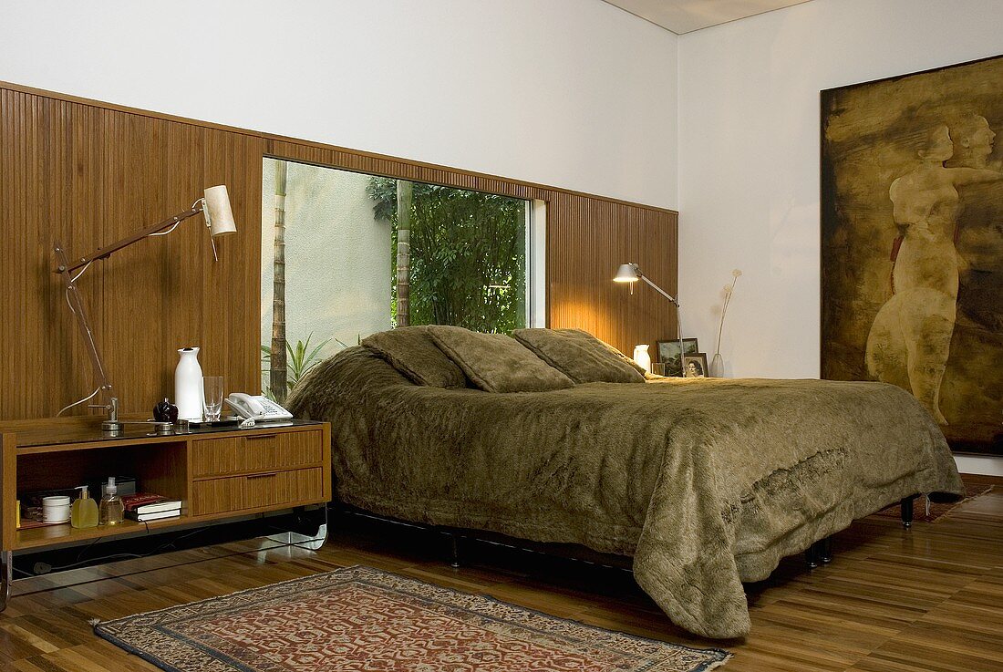 Doppelbett mit grüner Felltagesdecke vor Fenster und Nachttisch vor Holzwand aus gleichen Holz