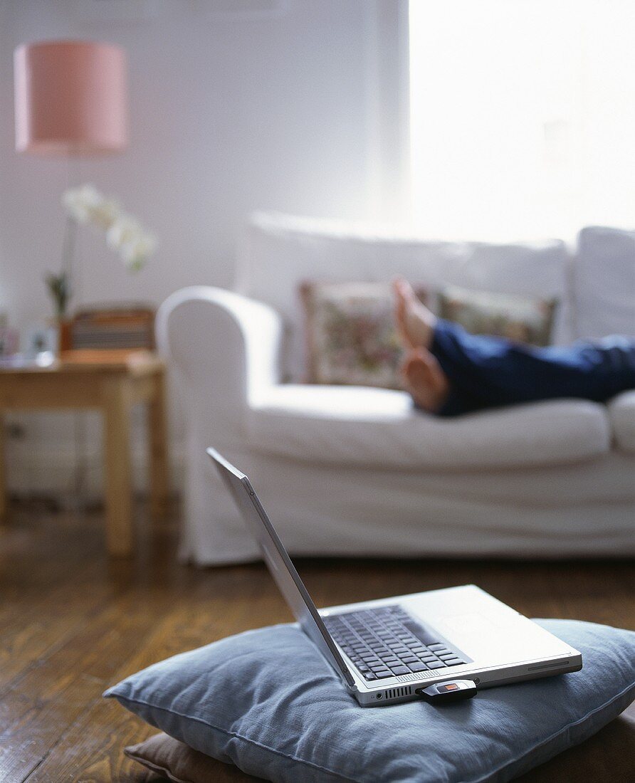 Laptop auf Kissen am Boden eines Wohnzimmers, im Hintergrund Frau auf Sofa liegend
