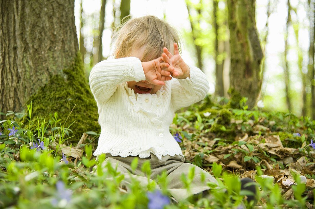 Kleinkind auf Waldboden sitzend, versteckt Gesicht mit Händen
