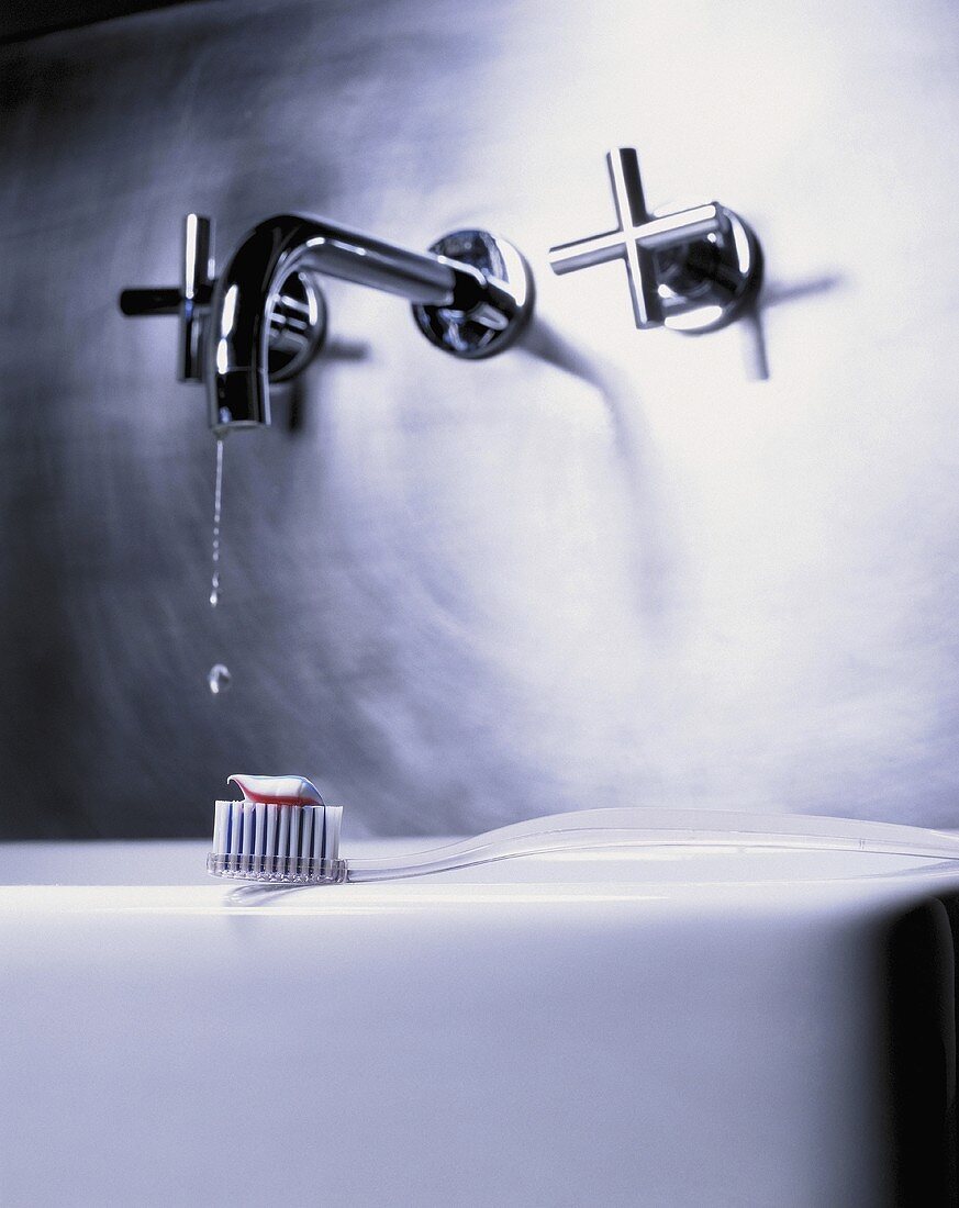 Zahnbürste mit Zahnpasta liegt auf dem Rand eines Waschbeckens mit moderner Armatur & tropfendem Wasserhahn