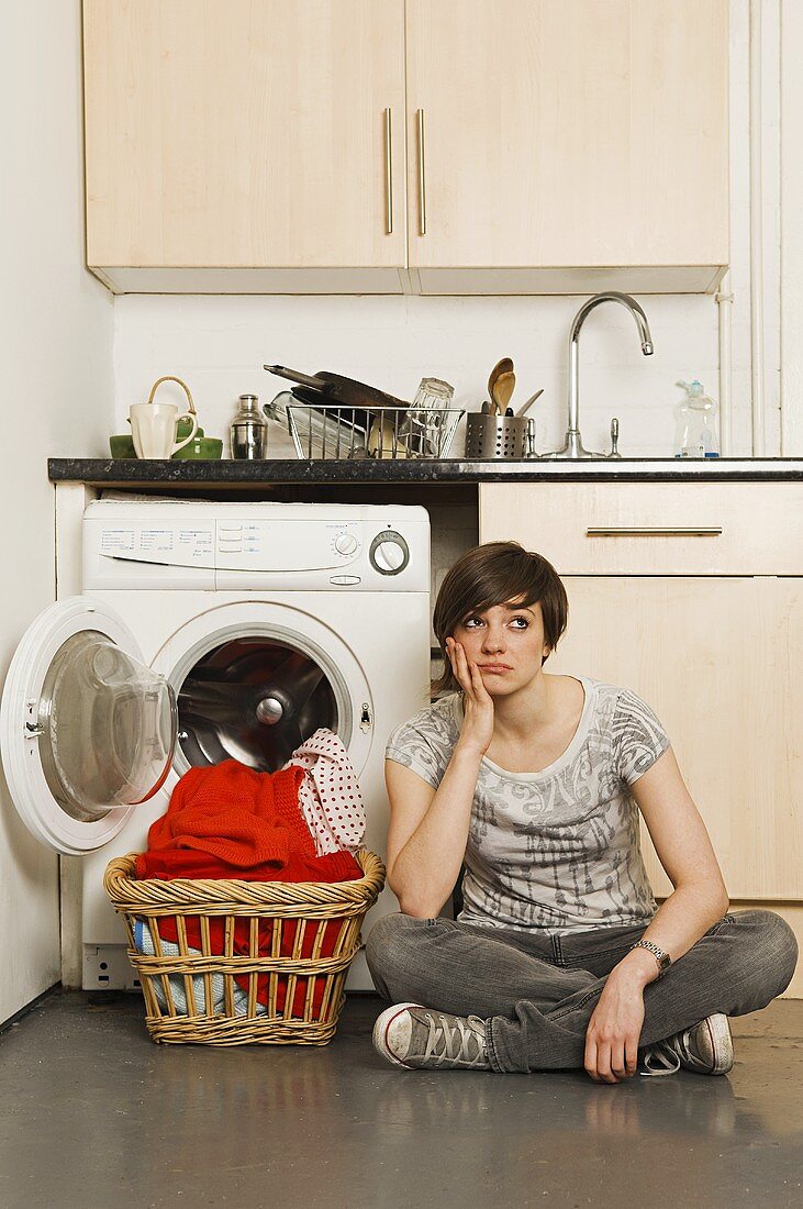 Gelangweilte junge Frau sitzt im Schneidersitz mit Wäschekorb vor Waschmaschine