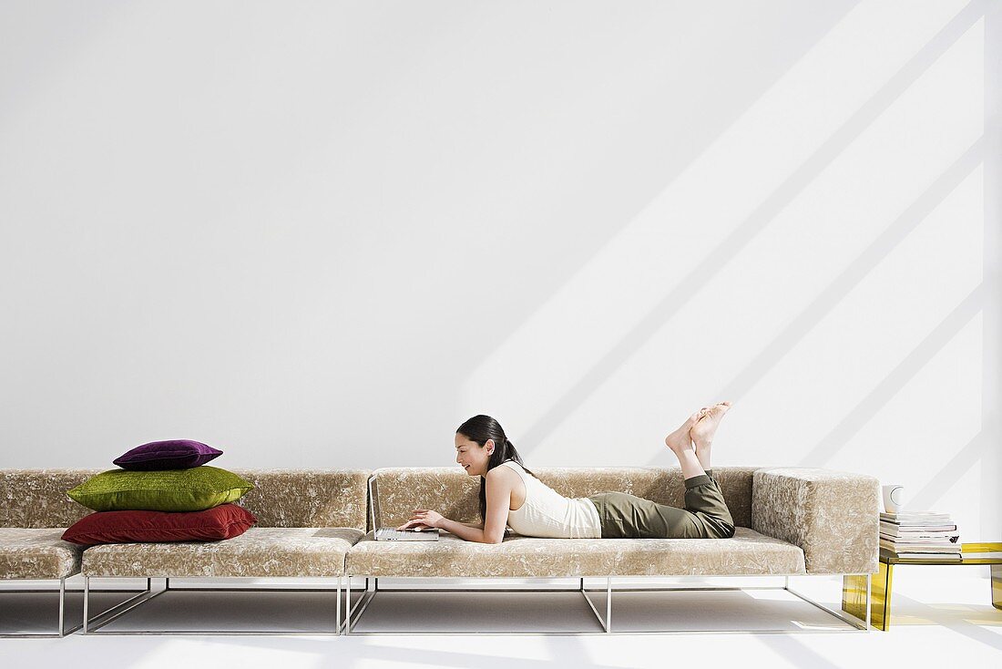 Junge Frau beim Lesen auf langgezogenem Sofa mit darauf gestapelten Samtkissen