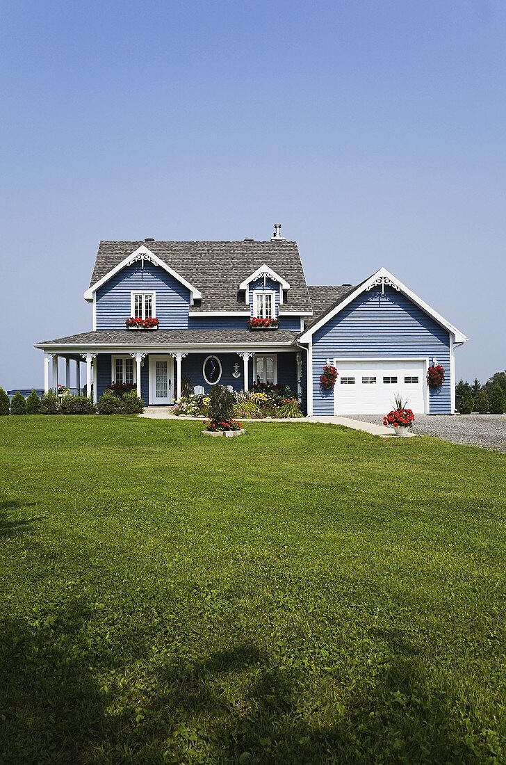 Pastellfarbenes, holzverkleidetes Haus mit Veranda und breitem Garagentor; davor eine große Wiese
