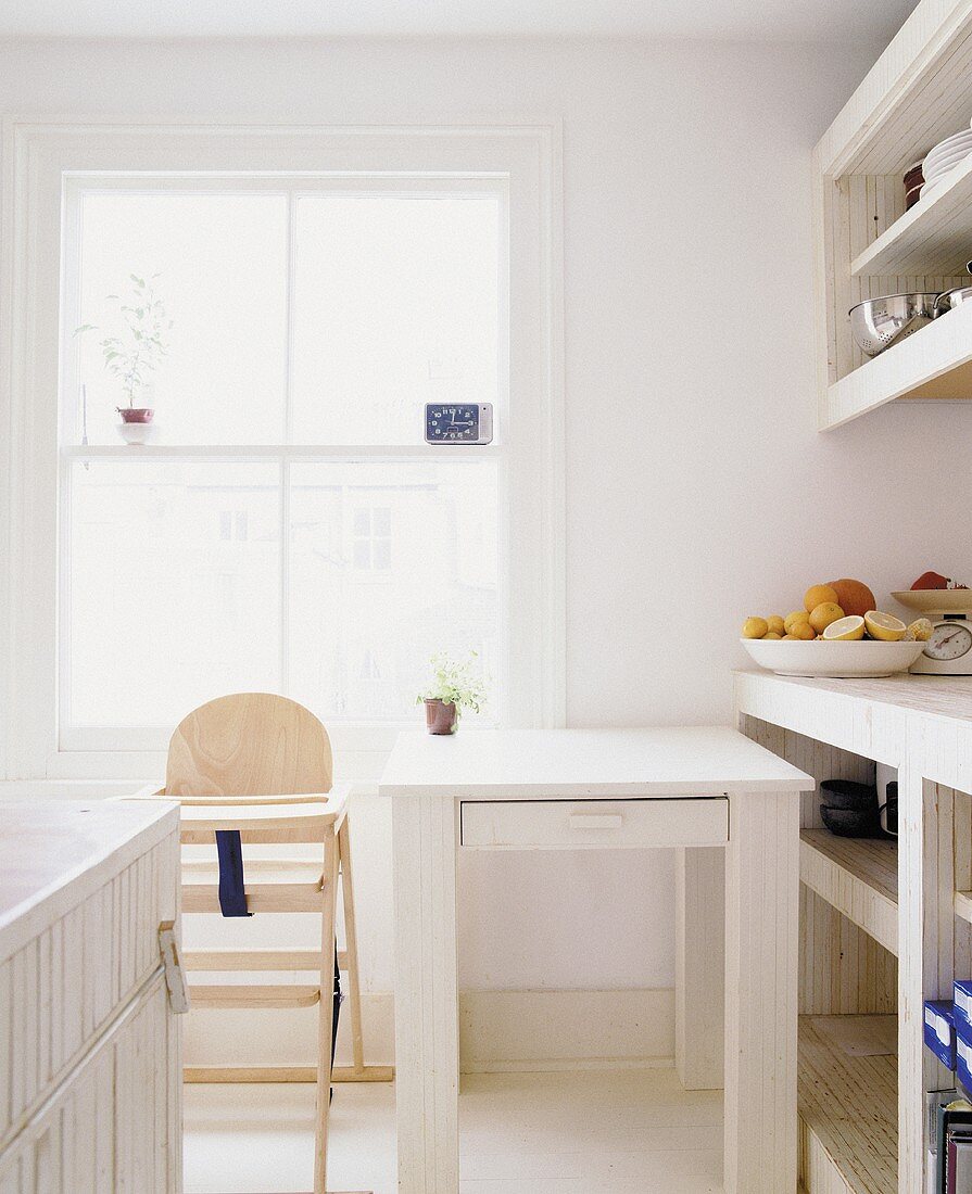 weiße, helle Küche mit Kinderhochstuhl und schlichtem Tisch am Fenster