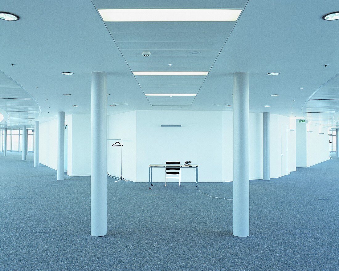 Weitläufiger, leerer Raum mit grauem Teppichboden, Säulen und einem zentralen Arbeitsplatz