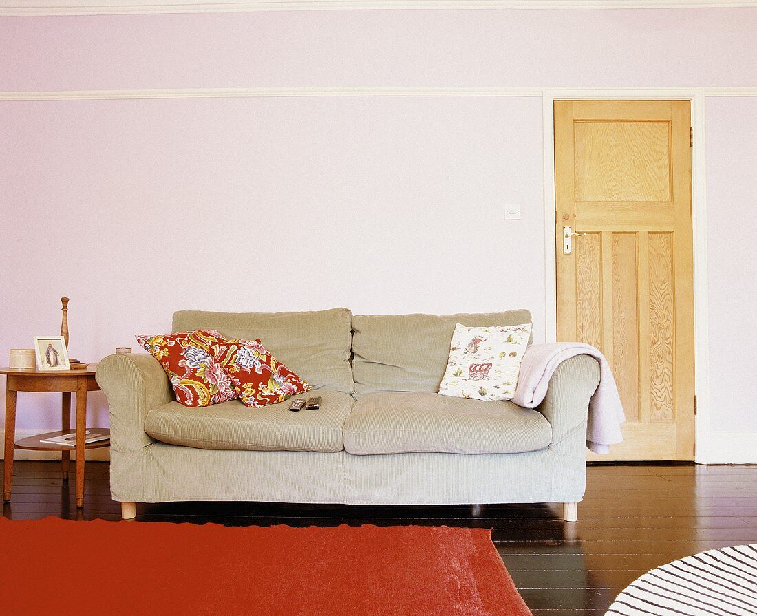 Schlichtes Wohnzimmer mit dunklem Dielenboden, orangefarbenem Teppich und einer einfachen Couch mit blumengemusterten Zierkissen