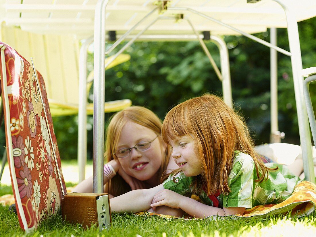 Zwei Mädchen liegen unter Gartentisch und hören Radio