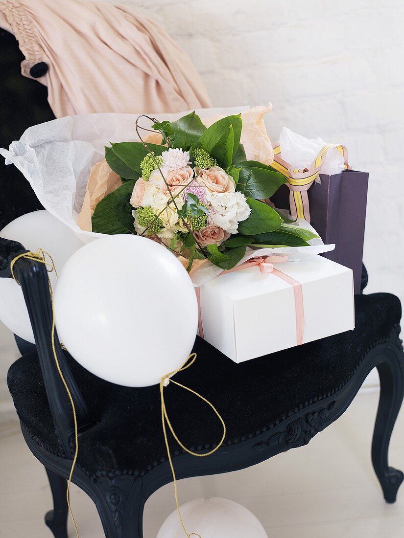 Romantischer Blumenstrauss, Geschenkpaket und weiße Luftballons auf schwarzem Barockstuhl