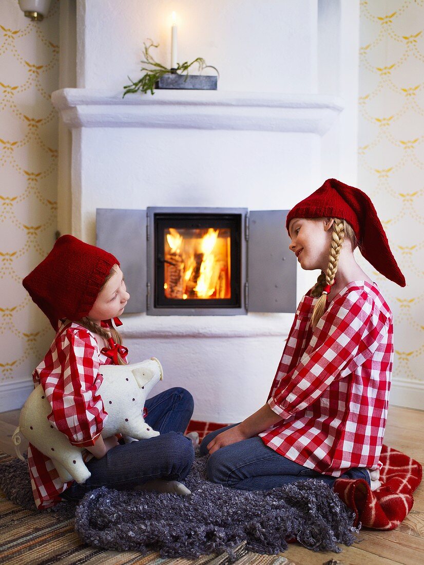 Zwei Mädchen mit Weihnachtsmützen und rot-weiss karierten Hemden vor dem Kaminfeuer