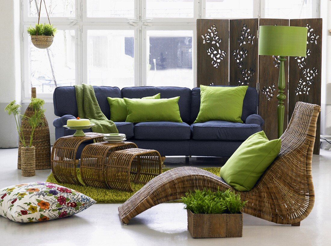 Wohnzimmer mit blauem Sofa, Korbmöbeln, grünen Kissen und einem Paravent mit Schnitzereien vor großer Fensterfront
