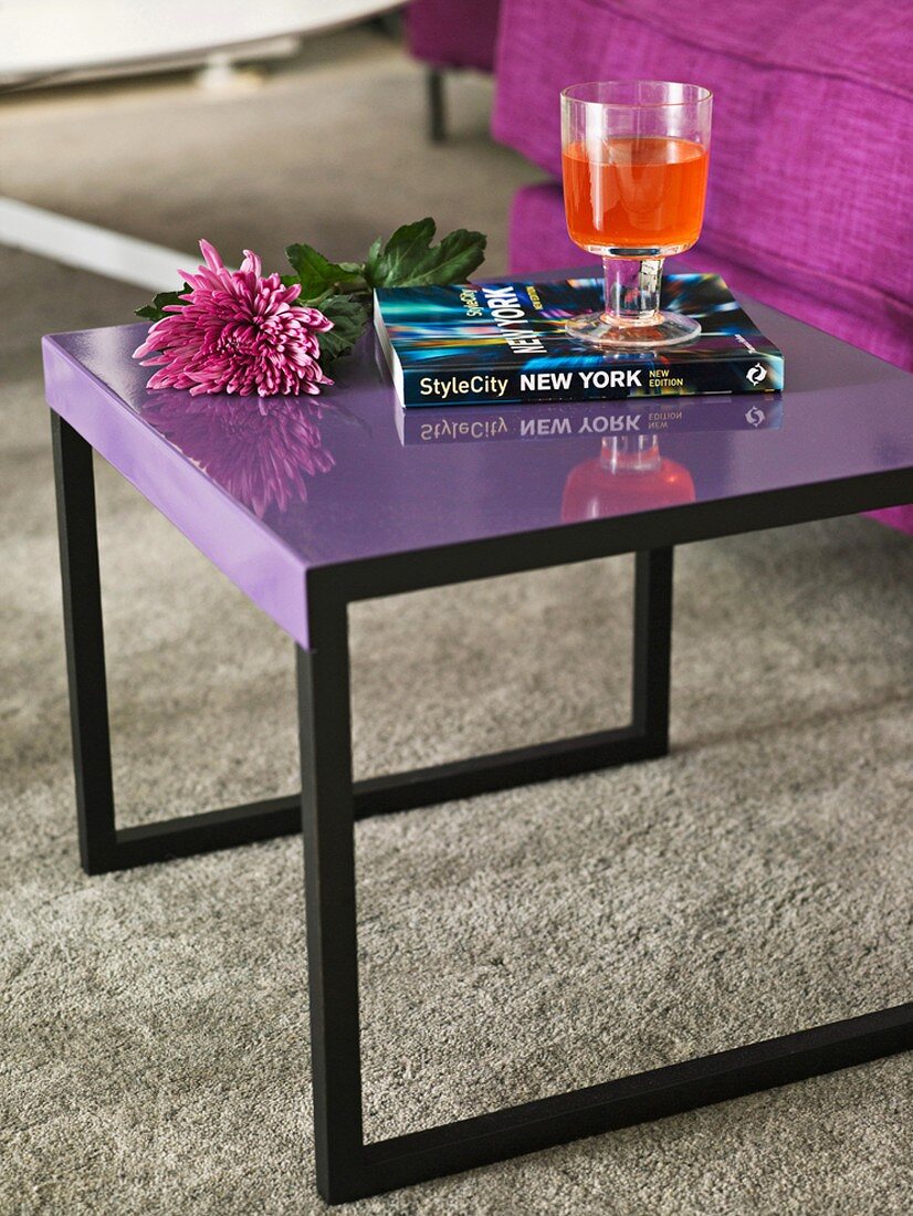 Lila Beistelltisch mit Buch, Blume und Drink vor violetter Couch