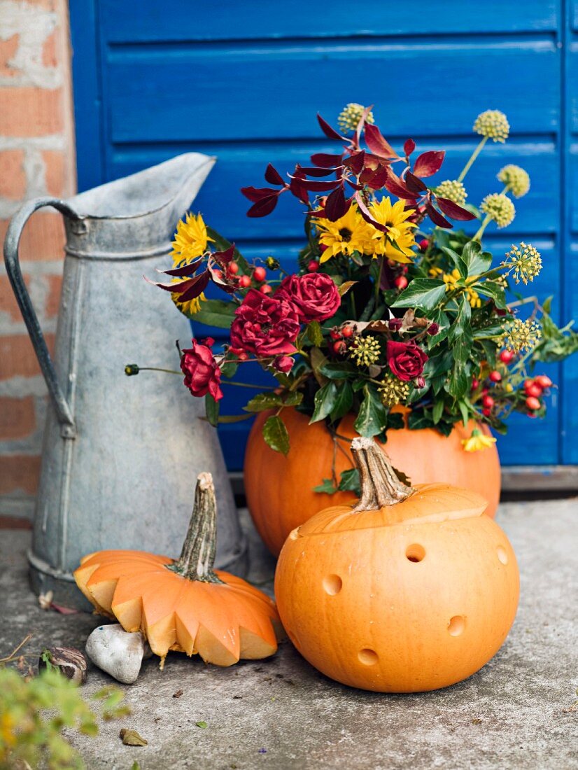 Metal jug, pumpkins and flower arrangement in front of house
