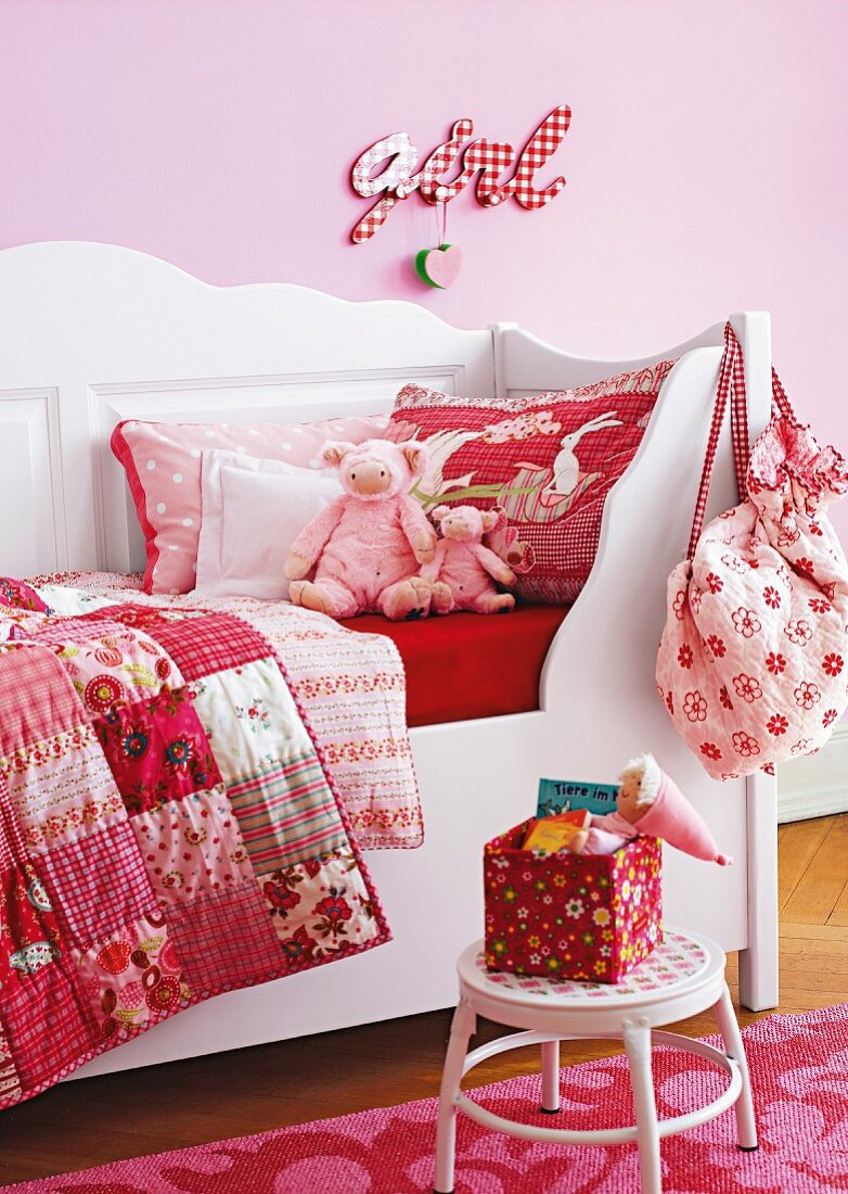 Weisses Kinderbett mit Kissen, Patchworkdecken in roten Tönen vor rosa Wand